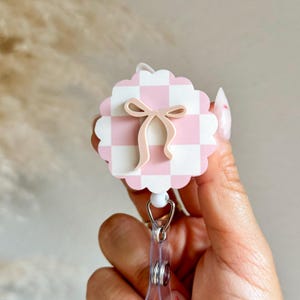 Pu&ograve; includere: Un portabadge a scacchi rosa e bianchi con un fiocco rosa chiaro. Il portabadge ha un bordo smerlato e una clip di plastica trasparente. Il design &egrave; un accessorio carino e femminile.