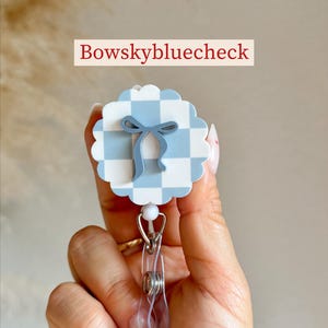 Pu&ograve; includere: Un portabadge a motivo gingham bianco e azzurro con un design a fiocco azzurro. Il portabadge ha una clip in plastica trasparente e un cordino retrattile in metallo. Il testo "Bowskybluecheck" &egrave; visualizzato.