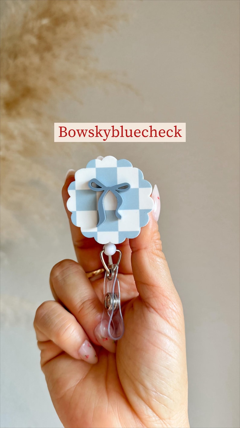 Pu&ograve; includere: Un portabadge a motivo gingham bianco e azzurro con un design a fiocco azzurro. Il portabadge ha una clip in plastica trasparente e un cordino retrattile in metallo. Il testo "Bowskybluecheck" &egrave; visualizzato.