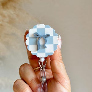 Pu&ograve; includere: Un portabadge a fantasia gingham azzurra e bianca con un design a fiocco blu. Il portabadge ha una clip in plastica trasparente e una perla bianca. Un accessorio carino.