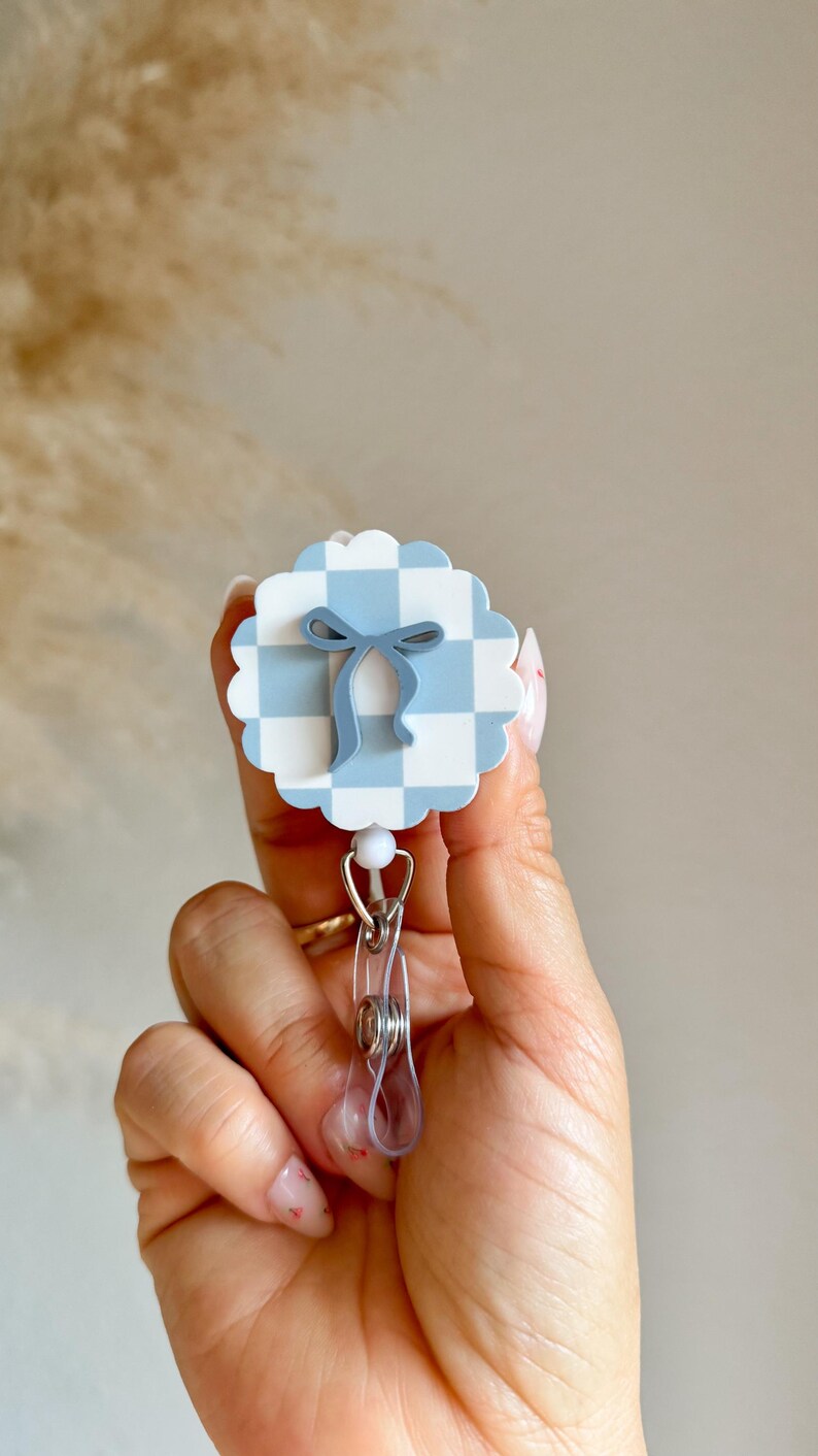 Pu&ograve; includere: Un portabadge a fantasia gingham azzurra e bianca con un design a fiocco blu. Il portabadge ha una clip in plastica trasparente e una perla bianca. Un accessorio carino.