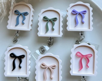 May include: Six badge reels with a white scalloped frame and a colorful bow design. The bows are in shades of blue, green, purple, black, pink, and peach. Each reel has a clear plastic strap and a metal clip.