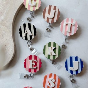 May include: A collection of colorful, striped badge reels with various letters. Each reel features a different color scheme, including pink, blue, green, and orange, with a white letter in the center. The reels are attached to clear plastic clips.