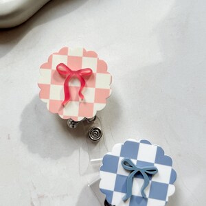 Pu&ograve; includere: Due porta badge con bordo smerlato. Uno ha un motivo a scacchi rosa e bianco con un fiocco rosa. L'altro ha un motivo a scacchi blu e bianco con un fiocco blu. Entrambi hanno un cinturino in plastica trasparente e una clip in metallo.