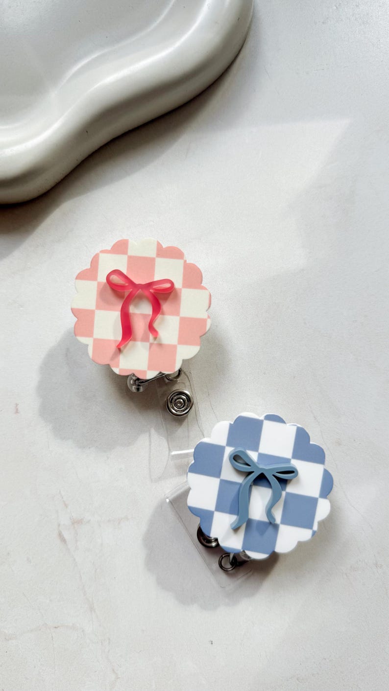 Pu&ograve; includere: Due porta badge con bordo smerlato. Uno ha un motivo a scacchi rosa e bianco con un fiocco rosa. L'altro ha un motivo a scacchi blu e bianco con un fiocco blu. Entrambi hanno un cinturino in plastica trasparente e una clip in metallo.