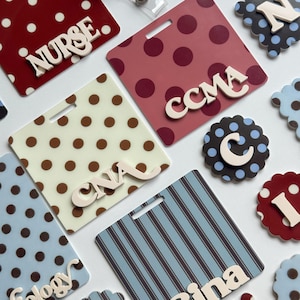 May include: A collection of square and scalloped acrylic badge reels featuring various patterns including polka dots and stripes. The square badges display text such as NURSE, CCMA, CNA, and Gina, while the scalloped pieces feature individual letters.