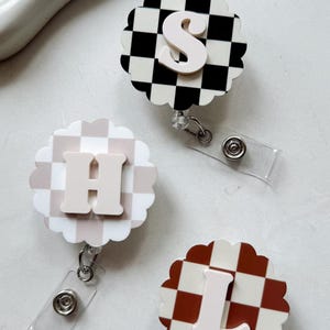 May include: Three badge reels with scalloped edges and checkered patterns. Each reel features a different letter: S in black and white, H in beige and white, and L in brown and white. The letters are in a contrasting color.