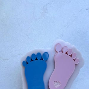 May include: A badge reel featuring two baby footprints, one blue and one pink, each with a heart cutout. The footprints are set against a white background. The reel has a white retractable cord and a metal clip.