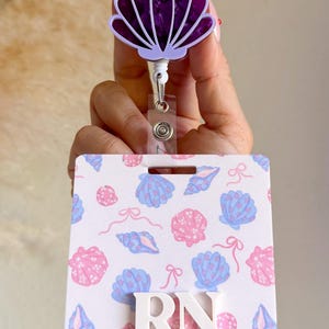 May include: A badge reel with a seashell design. The reel features a purple seashell-shaped top and a white retractable cord. The badge holder has a white background with pink and blue seashells and the letters "RN".