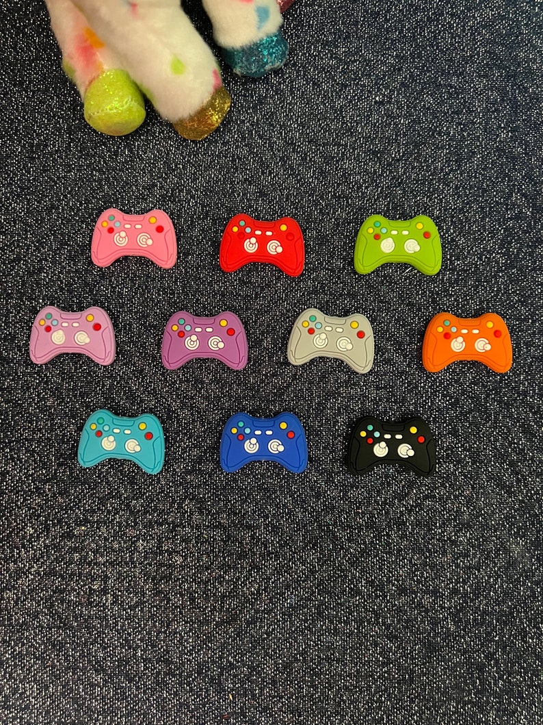 May include: Assortment of ten colorful plastic game controller charms. The charms are in various colors including pink, red, purple, gray, orange, blue, and black.