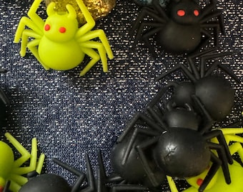 May include: A close-up of a group of black and green plastic spiders. The spiders have red eyes and are arranged on a blue denim background.