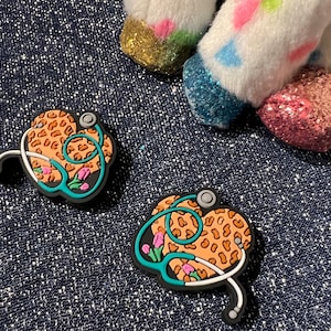 May include: Two black heart-shaped charms with a teal stethoscope design. The hearts are covered in a brown and orange leopard print with pink flowers.