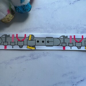 May include: A white grosgrain ribbon featuring a repeating graphic print of a grey tool belt. The belt design includes illustrations of hammers, pliers, screwdrivers, and yellow rulers, all arranged in a horizontal pattern along the length of the ribbon.