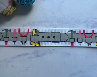 May include: A white grosgrain ribbon featuring a repeating graphic print of a grey tool belt. The belt design includes illustrations of hammers, pliers, screwdrivers, and yellow rulers, all arranged in a horizontal pattern along the length of the ribbon.