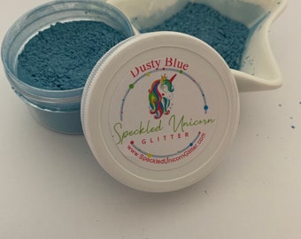 May include: A small jar of dusty blue glitter with a white lid. The lid has a colorful unicorn logo and the text "Speckled Unicorn Glitter" and "www.SpeckledUnicornGlitter.com".