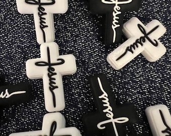 May include: A collection of small, cross-shaped charms in black and white. Each cross features the word Jesus written in a cursive script across the center. The charms are made of a solid material and are arranged in a scattered pattern.