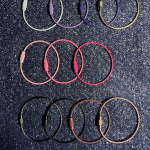 May include: A collection of keychains in various colors, including white, pink, red, purple, gold, and black. Each keychain features a circular wire design with a screw-on clasp. The keychains are arranged on a dark blue, glittery surface.