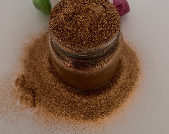 May include: A glass jar filled with fine, sparkly copper glitter. The glitter is scattered around the jar on a white surface.