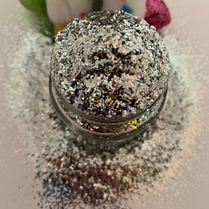 May include: A clear glass jar filled with silver glitter. The glitter is loose and has a slight iridescent sheen. The jar is sitting on a white surface.