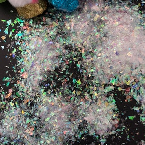 May include: Iridescent glitter flakes scattered on a black surface. The glitter reflects multiple colors, creating a shimmering effect. Small plush figures with glittery accents are visible in the background, suggesting a craft or party theme.