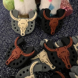 May include: A set of 10 black, brown, and gray plastic bull skull charms. The charms are shaped like a bull's skull with horns and have a hole in the top for attaching to a necklace or bracelet.