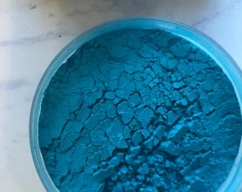 May include: A close-up of a small container filled with teal blue loose pigment powder. The powder has a cracked, dry texture.