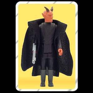 Op de afbeelding: Een roodhuidige action figure met hoorns en een zwarte cape, die een zilveren wapen vasthoudt. De figuur staat op een gele achtergrond.
