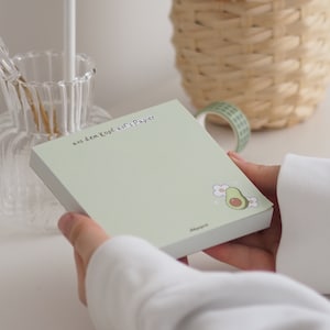 Könnte beinhalten: Ein grüner Notizblock mit dem Text "aus dem Kopf auf's Papier" und einer kleinen Avocado-Illustration. Der Notizblock wird von einer Hand gehalten.