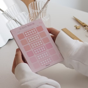 Könnte beinhalten: Ein rosa-weißes Notizbuch mit dem Text "JUST BREATHE YOU GOT THIS" und einer Wolkengrafik. Das Notizbuch hat ein Raster aus Quadraten mit weißen Punkten in jedem Quadrat. Das Notizbuch wird von einer Hand gehalten.