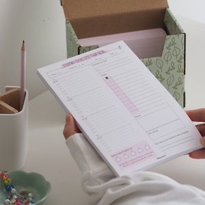 Könnte beinhalten: Ein weißer Notizblock mit rosa Akzenten und dem Text "STUDYING SUCKS LESS THAN FAILING" wird vor einer Box mit Blättermuster gehalten. Ein rosa Bleistift und eine kleine grüne Schale mit bunten Perlen sind ebenfalls zu sehen.