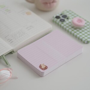 Könnte beinhalten: Ein rosa liniertes Notizbuch mit einer kleinen Igel-Illustration in der Ecke. Das Notizbuch liegt auf einer weißen Oberfläche, neben einem Planer, einer Kaffeetasse mit Lippenmotiv und einem Telefon mit grünem Vichy-Muster.