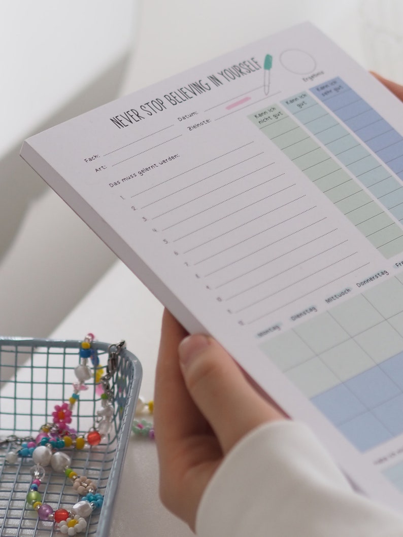 May include: A white notepad with the text "NEVER STOP BELIEVING IN YOURSELF" in black. The notepad has a section for "Fach", "Art", "Datum", "Zielnote", and a list of days of the week. There is a green pen on the notepad.