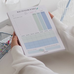 May include: A white notepad with the text "NEVER STOP BELIEVING IN YOURSELF" at the top. The notepad has a daily planner section with columns for "Date", "Reminder", "Kann ich nicht gut", "Kann ich gut", and "Kann ich sehr gut". The notepad also has a weekly planner section with columns for "Monday", "Tuesday", "Wednesday", "Thursday", "Friday", "Saturday", and "Sunday".