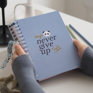 Könnte beinhalten: Ein blaues Notizbuch mit Spiralbindung mit dem Text "Hey! never give up" und einer Panda-Illustration.
