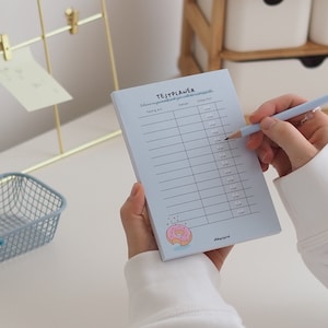 Könnte beinhalten: Ein hellblauer Notizblock mit einer Donut-Illustration und dem Text "TESTPLANER" oben. Der Notizblock hat Spalten mit den Bezeichnungen "Fach/Lern" und "Datum".