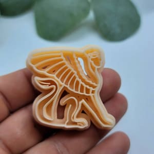 May include: A light orange plastic cookie cutter shaped like a sphinx with wings.