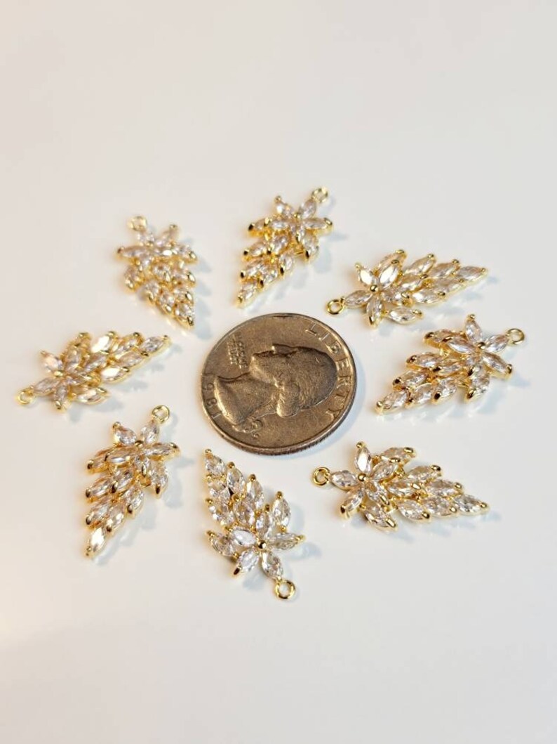 Puede incluir: Ocho abalorios en forma de hoja de color dorado con detalles de cristal transparente. Los abalorios est&aacute;n dispuestos en c&iacute;rculo alrededor de una moneda de veinticinco centavos de d&oacute;lar estadounidense.