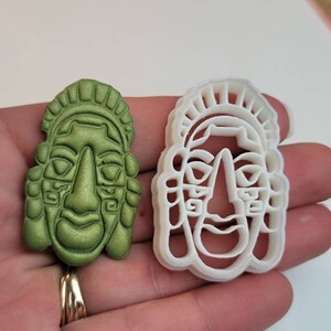 May include: A green, sculpted face and a white plastic face-shaped cookie cutter. The green face has detailed features, including a prominent nose, closed eyes, and a decorative headdress. The cookie cutter is a matching outline.
