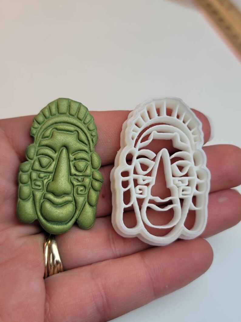 May include: A green, sculpted face and a white plastic face-shaped cookie cutter. The green face has detailed features, including a prominent nose, closed eyes, and a decorative headdress. The cookie cutter is a matching outline.
