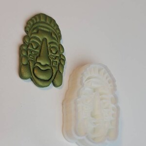 May include: An olive green, detailed mask and a white mask mold. The mask has a stylized face with a prominent nose, and detailed headwear. The mask mold is a translucent white, showing the same face design. The items are likely for crafting or art projects.