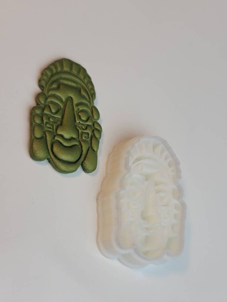 May include: An olive green, detailed mask and a white mask mold. The mask has a stylized face with a prominent nose, and detailed headwear. The mask mold is a translucent white, showing the same face design. The items are likely for crafting or art projects.
