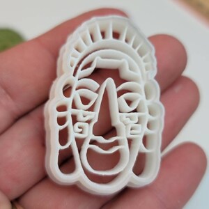 May include: A white plastic cookie cutter in the shape of a stylized face with a headdress. The cutter has a detailed design with facial features and decorative elements. The cutter is held in a hand.