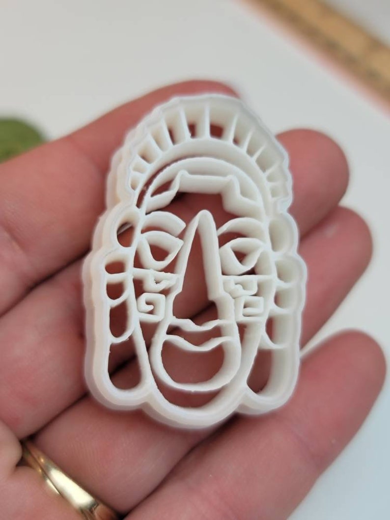May include: A white plastic cookie cutter in the shape of a stylized face with a headdress. The cutter has a detailed design with facial features and decorative elements. The cutter is held in a hand.