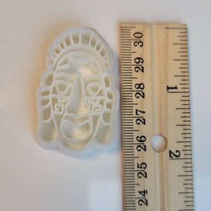 May include: A white plastic cookie cutter in the shape of a stylized face, approximately 3 inches tall. The face has a prominent nose, rectangular eyes, and a headdress. A wooden ruler is next to the cutter.