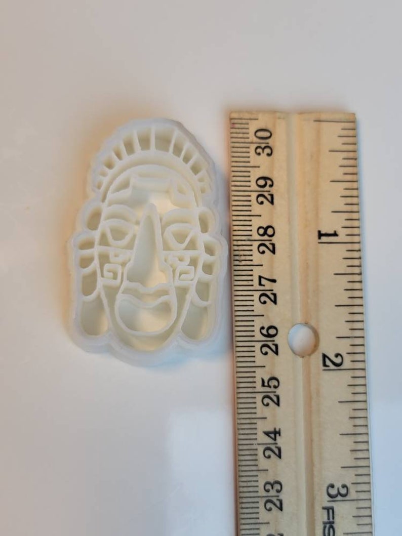 May include: A white plastic cookie cutter in the shape of a stylized face, approximately 3 inches tall. The face has a prominent nose, rectangular eyes, and a headdress. A wooden ruler is next to the cutter.