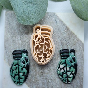 Puede incluir: Tres adornos de resina en forma de corazón. El adorno central es de color marrón claro y presenta un diseño de cerebro detallado. Los dos adornos de los lados son verdes y negros con un diseño de cerebro y un anillo negro.