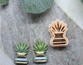 May include: Two green and gold succulent plant cookie cutters and a larger, plain, light peach succulent cookie cutter on a grey and white marble surface.