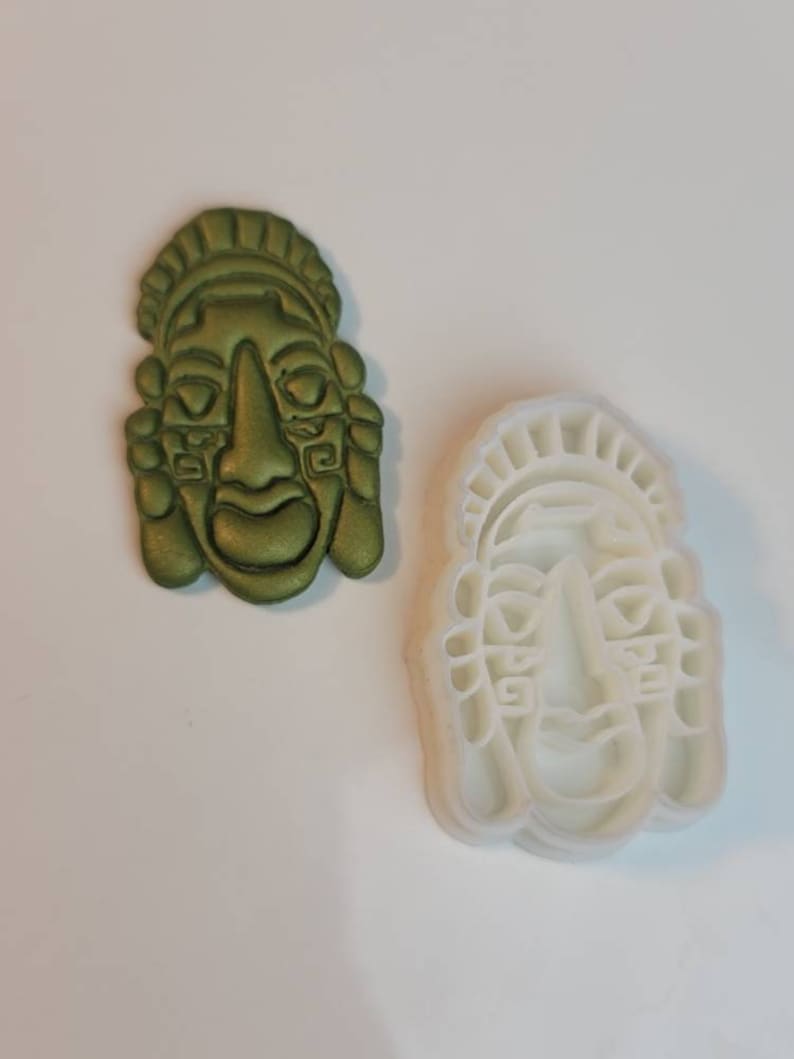 May include: A green, metallic-looking Aztec mask and a white plastic mask-shaped cookie cutter. The mask has detailed features, including a prominent nose, closed eyes, and ornate headwear. The cookie cutter is a reverse image of the mask.