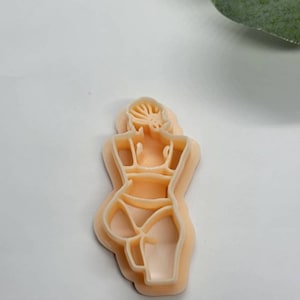 Puede incluir: Un cortador de galletas de color melocotón claro con forma de figura femenina, vista desde atrás. El cortador tiene líneas detalladas que definen los contornos del cuerpo, incluyendo el pelo, los hombros y las piernas. El diseño es simple y elegante.