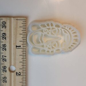 May include: A white plastic cookie cutter in the shape of a stylized face, approximately 2.5 inches long. The design includes facial features and decorative elements around the face. A wooden ruler is next to the cookie cutter.